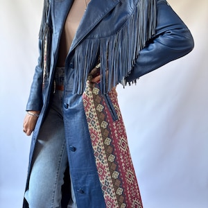 May include: A long blue leather coat featuring long fringe detailing across the chest and shoulders. The coat includes a button-front closure and vertical panels of woven fabric with a multi-colored geometric pattern in red, brown, and cream.