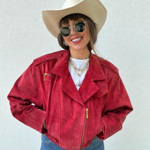 May include: A red suede jacket with gold zippers, paired with blue jeans and a cream-colored cowboy hat. The jacket has a classic biker style with a notched collar and a zippered pocket on the chest. The outfit is completed with round sunglasses and gold necklaces.