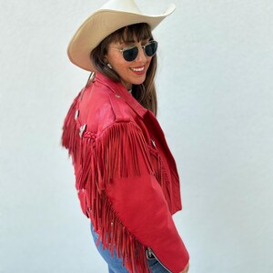 Op de afbeelding: Rode leren jas met franjes en zilveren accenten. De jas wordt gecombineerd met een cr&egrave;mekleurige cowboyhoed en een blauwe spijkerbroek. De jas heeft een ritssluiting.
