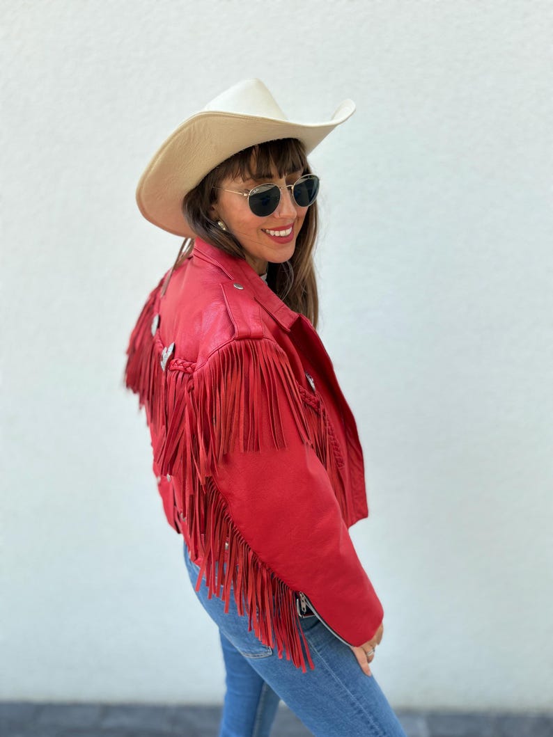 Op de afbeelding: Rode leren jas met franjes en zilveren accenten. De jas wordt gecombineerd met een cr&egrave;mekleurige cowboyhoed en een blauwe spijkerbroek. De jas heeft een ritssluiting.