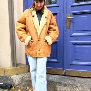 May include: A tan shearling coat with a cream-colored interior and trim. The coat has a button closure and two front pockets. The person is wearing light blue jeans and a white beanie. The background features a blue door.