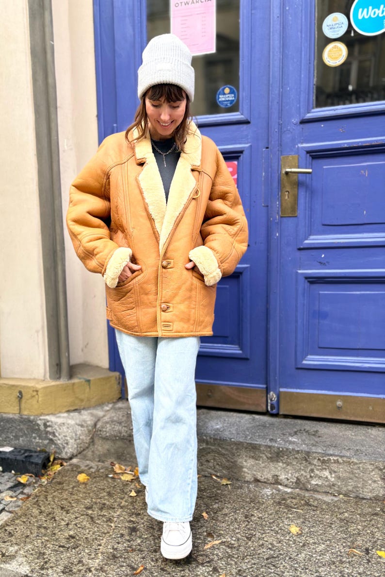May include: A tan shearling coat with a cream-colored interior and trim. The coat has a button closure and two front pockets. The person is wearing light blue jeans and a white beanie. The background features a blue door.