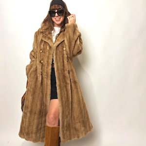 Puede incluir: Abrigo largo de piel sintética marrón claro con grandes botones dorados. El abrigo se lleva con una diadema negra, gafas de sol y botas de ante marrón. El abrigo tiene un estilo clásico y vintage.