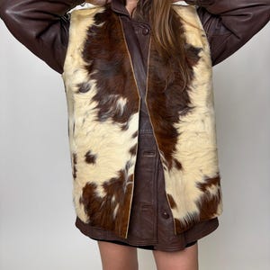 May include: A brown leather jacket with a cowhide vest. The vest has a cream-colored background with brown patches. The jacket has brown leather sleeves and a brown leather collar. The vest is buttoned up.
