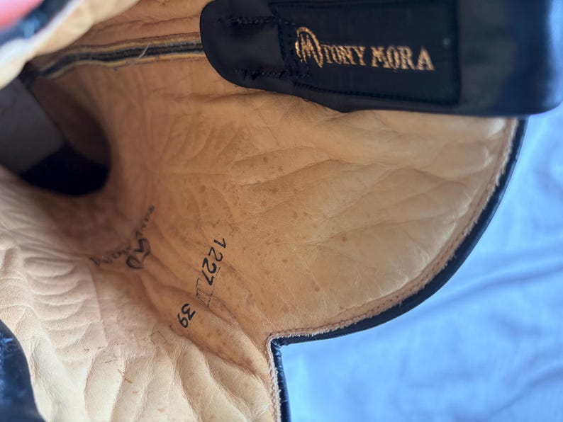 Puede incluir: Vista interior de una bota de cuero negro con una plantilla beige. La bota presenta el nombre de la marca "TONY MORA" en letras doradas. La plantilla muestra los n&uacute;meros "50", "1227" y "39".