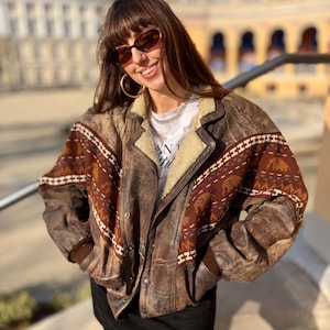 Puede incluir: Una chaqueta de cuero marrón vintage con un diseño estampado en los hombros y las mangas. La chaqueta tiene un forro de borreguillo color crema alrededor del cuello y una parte delantera abotonada. La chaqueta se usa con una camiseta blanca y una falda negra.