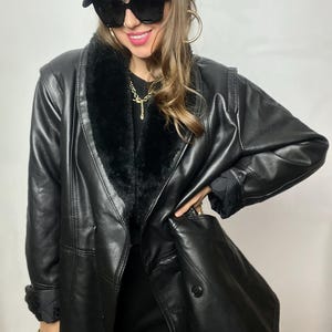 May include: A black faux leather jacket with a fur collar and a double-breasted closure. The jacket has a long, fitted silhouette and is worn over a black dress.