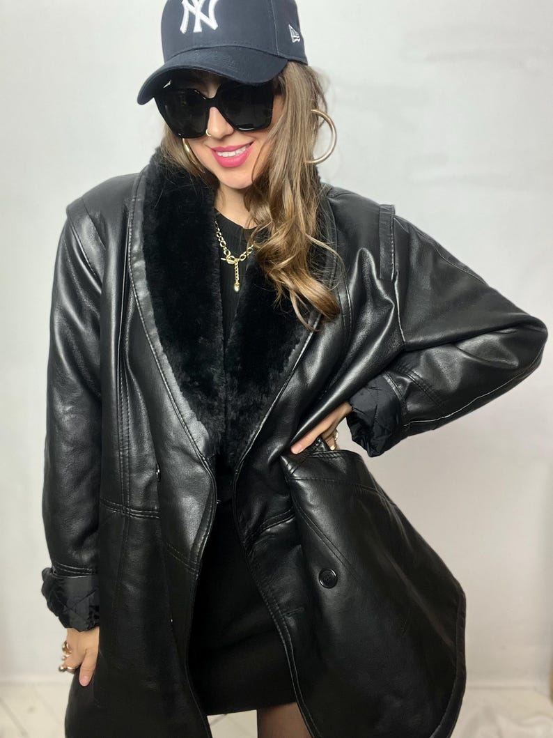 May include: A black faux leather jacket with a fur collar and a double-breasted closure. The jacket has a long, fitted silhouette and is worn over a black dress.
