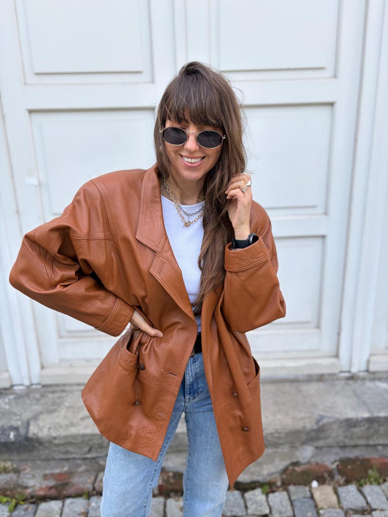 May include: A cognac-coloured leather blazer featuring a double-breasted front with dark buttons, wide lapels, and long sleeves. The jacket is worn over a plain white t-shirt and light blue denim jeans, paired with oval-shaped dark sunglasses.