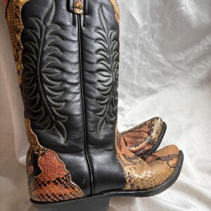Puede incluir: Botas de vaquero negras y marrones con estampado de piel de serpiente. Las botas tienen una punta afilada y una ca&ntilde;a alta. La ca&ntilde;a es negra con costuras decorativas. El estampado de piel de serpiente est&aacute; en la pala y la parte superior de la bota.