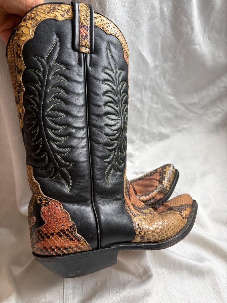 Puede incluir: Botas de vaquero negras y marrones con estampado de piel de serpiente. Las botas tienen una punta afilada y una ca&ntilde;a alta. La ca&ntilde;a es negra con costuras decorativas. El estampado de piel de serpiente est&aacute; en la pala y la parte superior de la bota.