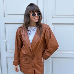 May include: A cognac-coloured leather blazer featuring a double-breasted front with two visible buttons, wide lapels, and long sleeves. The jacket is worn over a plain white top and paired with light blue denim jeans, complemented by oval sunglasses.