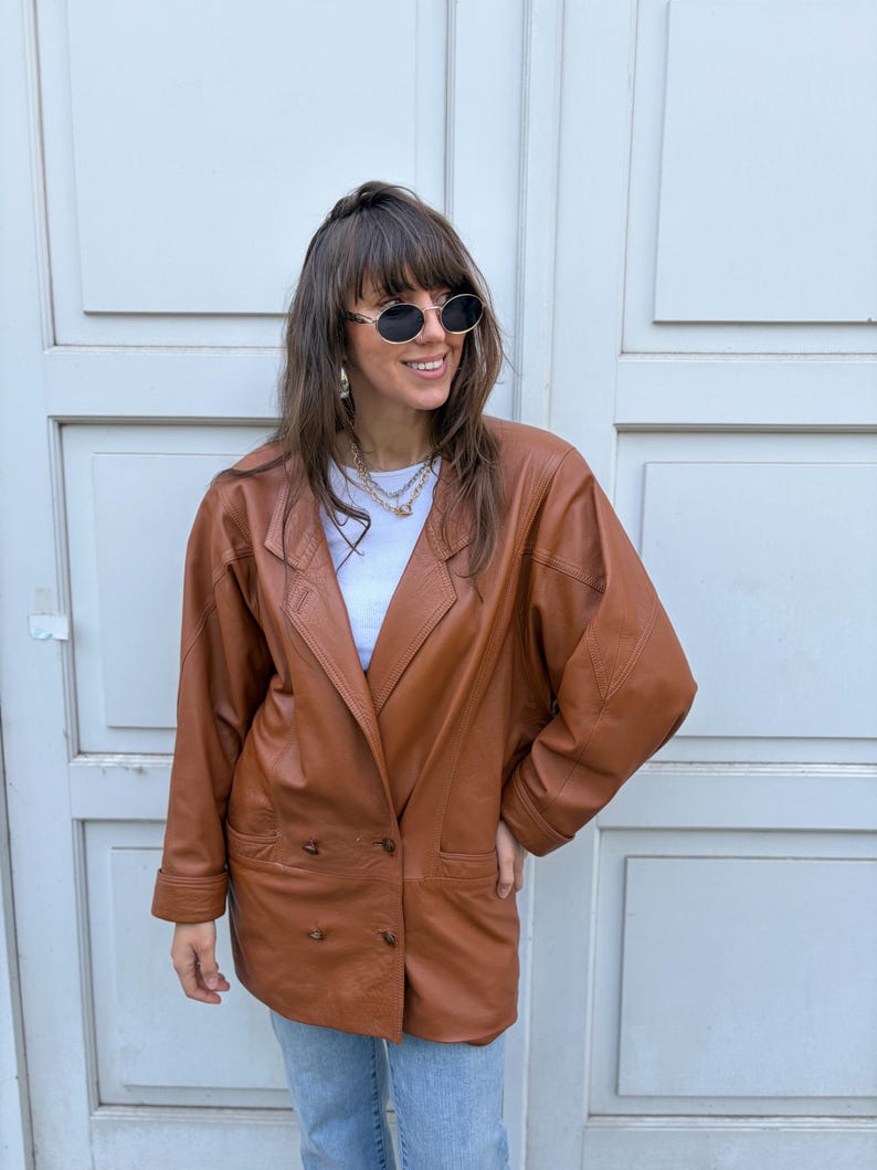 May include: A cognac-coloured leather blazer featuring a double-breasted front with two visible buttons, wide lapels, and long sleeves. The jacket is worn over a plain white top and paired with light blue denim jeans, complemented by oval sunglasses.