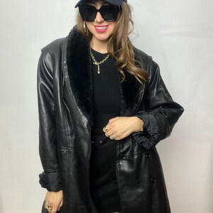 May include: A black faux leather jacket with a fur collar and a double breasted closure. The jacket has a belt at the waist and a New York Yankees baseball cap is worn on top of the head.