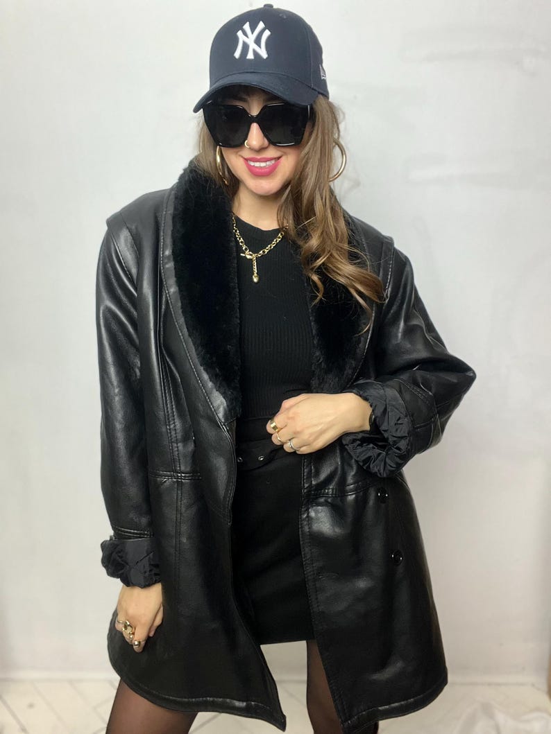 May include: A black faux leather jacket with a fur collar and a double breasted closure. The jacket has a belt at the waist and a New York Yankees baseball cap is worn on top of the head.