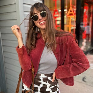 May include: A woman wearing a red suede jacket, gray top, and a cow print skirt. She is wearing gold-rimmed sunglasses and large hoop earrings. A brown leather handbag is slung over her shoulder. The background includes a storefront with red neon lettering.