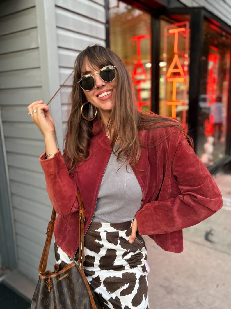 May include: A woman wearing a red suede jacket, gray top, and a cow print skirt. She is wearing gold-rimmed sunglasses and large hoop earrings. A brown leather handbag is slung over her shoulder. The background includes a storefront with red neon lettering.