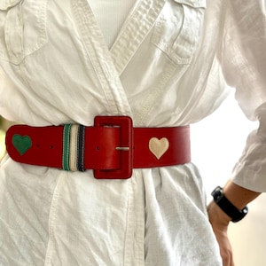 May include: A red leather belt with a large square buckle. The belt features a green heart and a cream heart design. A section of the belt has green, black, and white stripes. The belt is worn over a white shirt.