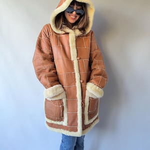 Può includere: Un cappotto in pelle color cuoio con un design a patchwork e cuciture bianche a vista, un cappuccio foderato in pelliccia sintetica color crema, due tasche anteriori con bordo in pelliccia sintetica e tre grandi bottoni rotondi centrali.
