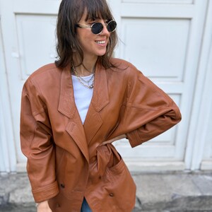 May include: A cognac-coloured leather blazer featuring a double-breasted front with visible buttons, wide lapels, and long sleeves. The jacket has a structured shoulder design and two front pockets, worn over a plain white top and light blue jeans.