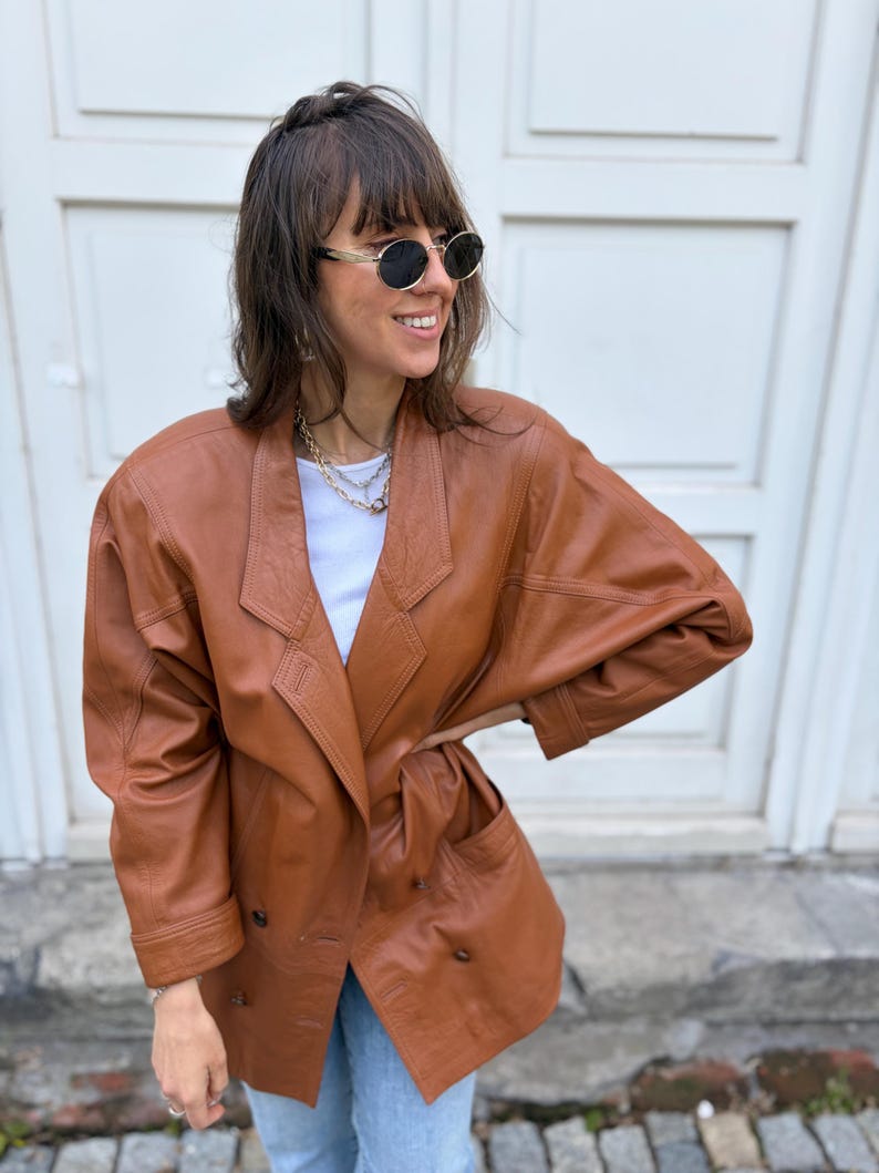 May include: A cognac-coloured leather blazer featuring a double-breasted front with visible buttons, wide lapels, and long sleeves. The jacket has a structured shoulder design and two front pockets, worn over a plain white top and light blue jeans.