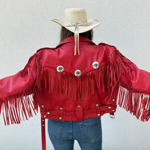 Op de afbeelding: Een felrode leren jas met lange franjes op de rug en mouwen. De jas heeft zilverkleurige metalen knopen en een riem. Een cr&egrave;mekleurige cowboyhoed met een gevlochten band en kinriem is ook zichtbaar.