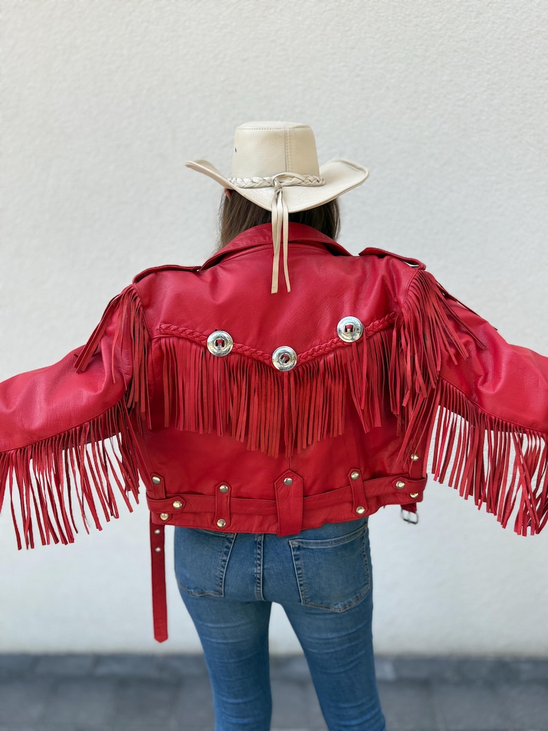Op de afbeelding: Een felrode leren jas met lange franjes op de rug en mouwen. De jas heeft zilverkleurige metalen knopen en een riem. Een cr&egrave;mekleurige cowboyhoed met een gevlochten band en kinriem is ook zichtbaar.