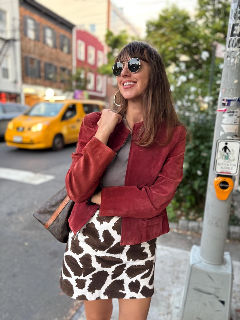 May include: A burgundy suede jacket and a brown and white patterned skirt. The jacket has a round neckline and long sleeves. The skirt has a high waist and a mini length. The woman is wearing sunglasses and carrying a brown handbag.