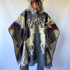 May include: A charcoal grey poncho featuring a cream-coloured graphic of two facing reindeer with large antlers. The garment has a faux fur collar, a single button closure at the neck, and a geometric pattern along the edges with a fringed hem.