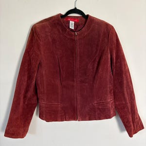 May include: A burgundy suede jacket with a zip-up front and two front pockets. The jacket is hanging on a black hanger. The label reads "AK Anne Klein".