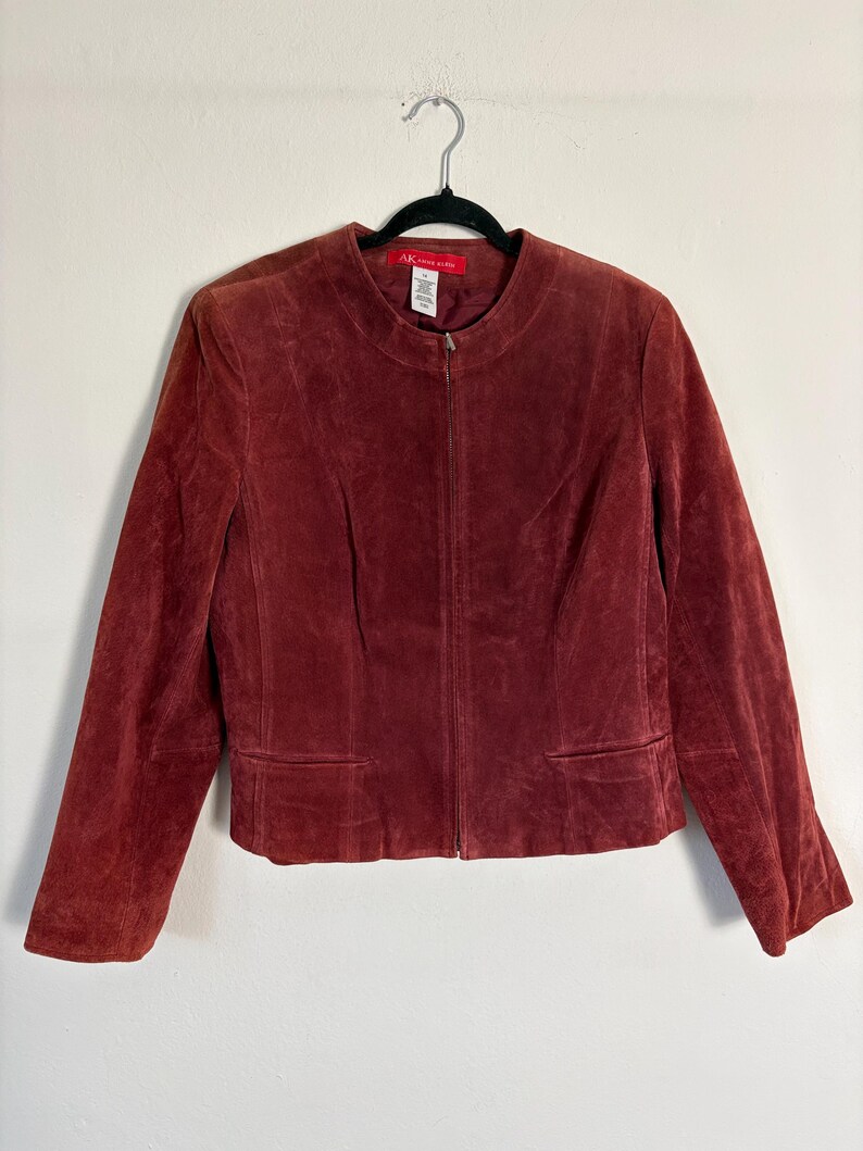 May include: A burgundy suede jacket with a zip-up front and two front pockets. The jacket is hanging on a black hanger. The label reads "AK Anne Klein".