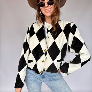 Puede incluir: Una chaqueta de manga larga con un patrón de rombos en blanco y negro. La chaqueta presenta un cuello redondo sin solapas, cierre frontal con botones dorados en forma de pirámide y dos pequeños bolsillos ribeteados en la cintura.