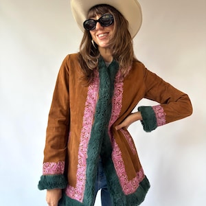 Könnte beinhalten: Eine braune Wildlederjacke mit dunkelgrünem Kunstfellbesatz an Kragen, Ärmelbündchen und Saum. Die Jacke ist mit vertikalen Bahnen aus rosa Blumenstickerei entlang der vorderen Öffnung und an den Ärmelbündchen verziert.