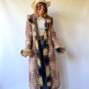 May include: A long, brown leopard-print coat featuring a thick, dark-tipped faux fur trim along the hood, front opening, and sleeve cuffs. The coat has a visible seam construction and is worn over a light grey top, blue denim jeans, and a brown belt.