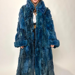 May include: A long, plush, deep teal blue faux fur coat featuring a high collar, long sleeves with wide cuffs, and a button-front closure. The coat has a thick, textured appearance with varying shades of blue throughout the dense, shaggy material.
