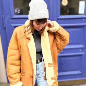 May include: A light brown shearling jacket with a cream-colored interior and trim. The jacket has a button closure, pockets, and a classic collar. A gray beanie with a small patch is worn on the head.