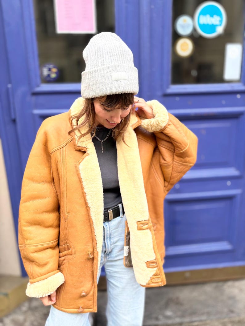 May include: A light brown shearling jacket with a cream-colored interior and trim. The jacket has a button closure, pockets, and a classic collar. A gray beanie with a small patch is worn on the head.