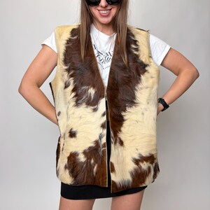 May include: A vintage cowhide vest with a cream and brown pattern. The vest has a V-neck and a zipper closure. The model is wearing a white t-shirt, black sunglasses, and a brown beret.