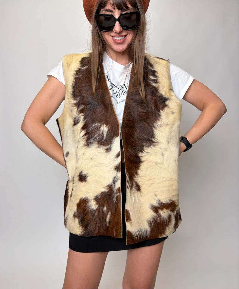 May include: A vintage cowhide vest with a cream and brown pattern. The vest has a V-neck and a zipper closure. The model is wearing a white t-shirt, black sunglasses, and a brown beret.