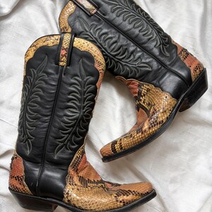Puede incluir: Un par de botas de vaquero negras con detalles de piel de serpiente en tonos marr&oacute;n, naranja y amarillo. Las botas presentan costuras decorativas y un dise&ntilde;o Western cl&aacute;sico.