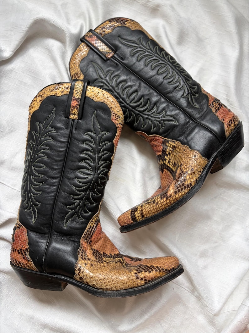 Puede incluir: Un par de botas de vaquero negras con detalles de piel de serpiente en tonos marr&oacute;n, naranja y amarillo. Las botas presentan costuras decorativas y un dise&ntilde;o Western cl&aacute;sico.