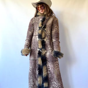 Puede incluir: Un abrigo largo de color marrón con estampado de leopardo que presenta un grueso borde de piel sintética con puntas oscuras a lo largo de la abertura frontal, el cuello y los puños. El abrigo se cierra con tres botones de palanca y un sombrero.