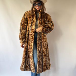 May include: A long, knee-length coat featuring a brown and tan animal print pattern with vertical dark brown stripes. The coat has long sleeves, a high collar, and a button-front closure. It is worn over a grey top, blue denim jeans, and brown boots.