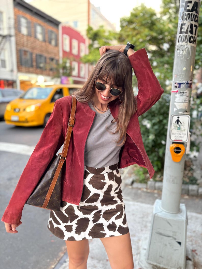 May include: A burgundy suede jacket, gray t-shirt, and a brown and white animal print mini skirt. A brown leather shoulder bag is worn over the shoulder. The person is wearing sunglasses and a watch.
