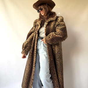 May include: A long, full-length faux fur coat featuring a brown and black leopard print pattern. The coat is worn over a light gray top, light blue denim jeans with a black belt, and is paired with a brown wide-brimmed hat and dark sunglasses.