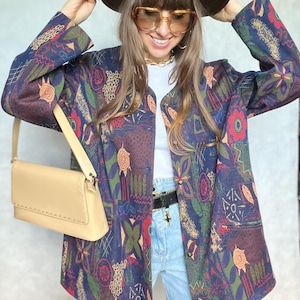 May include: A woman wearing a brown hat, a white shirt, blue jeans, and a colorful patterned jacket. She is carrying a light brown shoulder bag.
