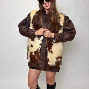 May include: A brown leather jacket with a cowhide vest, a brown beret, and black sunglasses. The vest features a cream and brown pattern. The person is wearing black boots. The outfit is a mix of textures and colors.