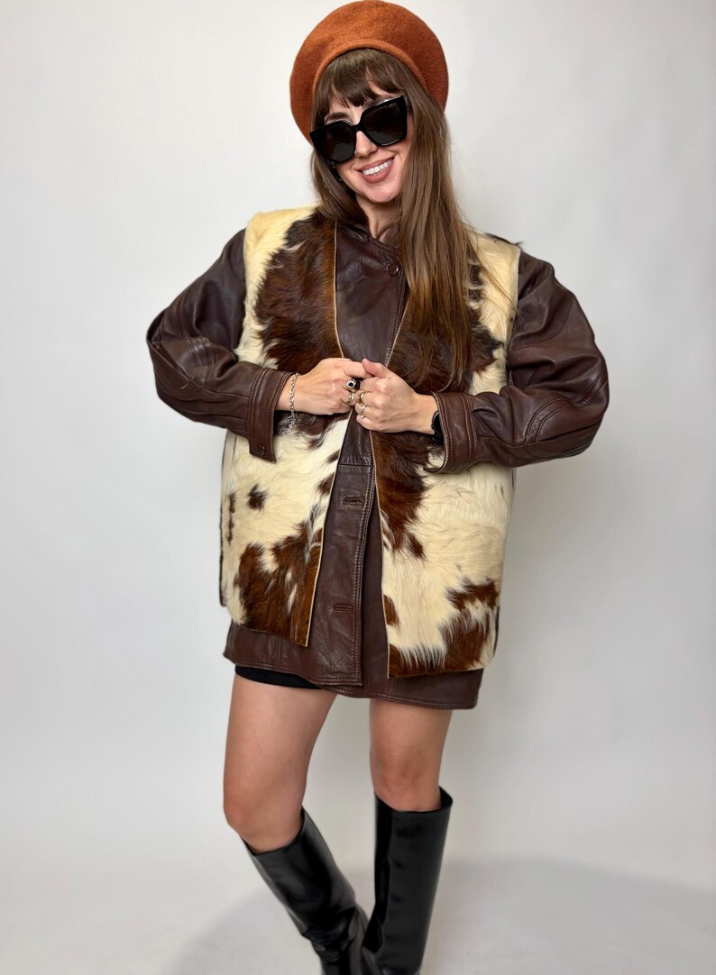 May include: A brown leather jacket with a cowhide vest, a brown beret, and black sunglasses. The vest features a cream and brown pattern. The person is wearing black boots. The outfit is a mix of textures and colors.