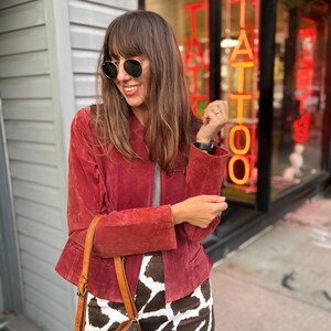 May include: A woman wearing a red suede jacket, round sunglasses, and a brown and white patterned skirt. She is carrying a brown leather bucket bag with a drawstring closure. The background includes a storefront with the word "TATTOO" in neon.