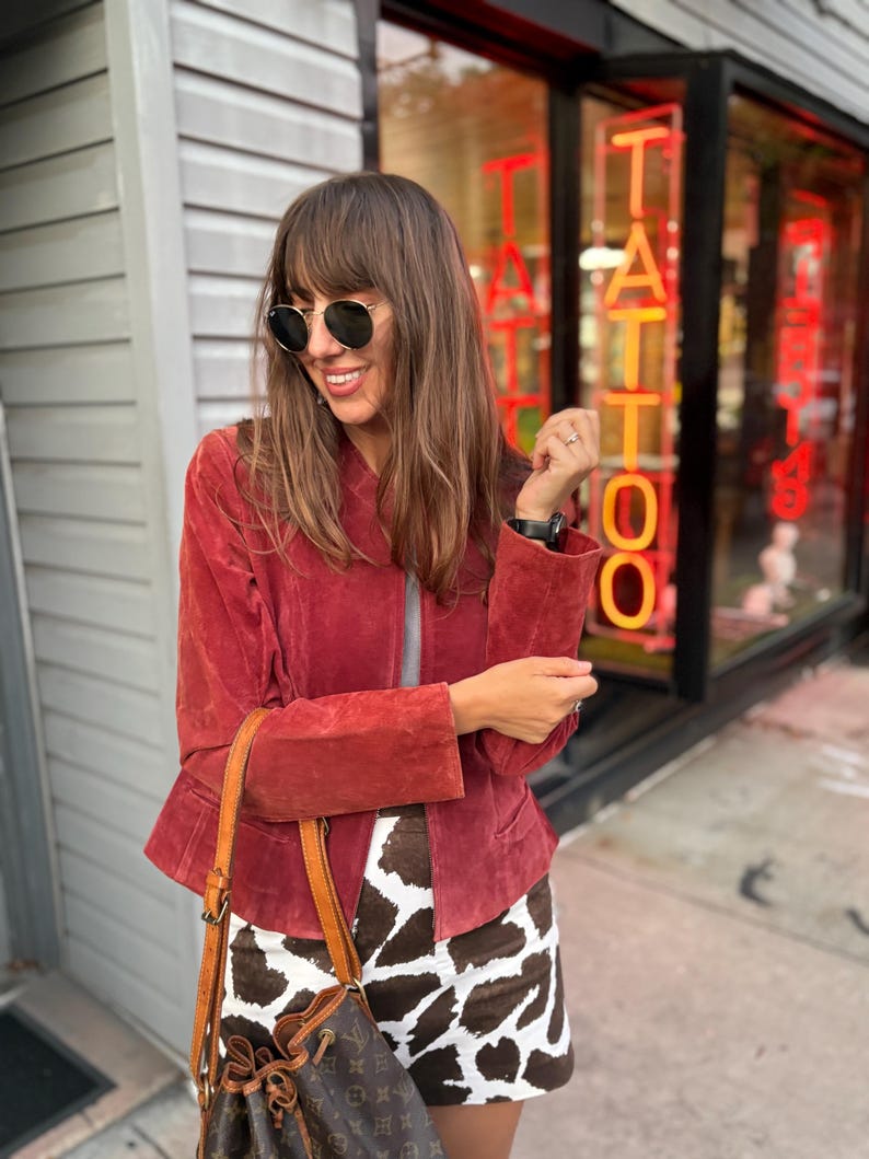 May include: A woman wearing a red suede jacket, round sunglasses, and a brown and white patterned skirt. She is carrying a brown leather bucket bag with a drawstring closure. The background includes a storefront with the word "TATTOO" in neon.
